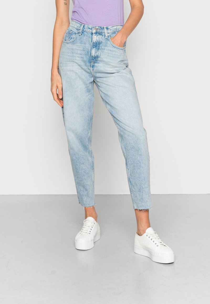 Person som bär ljusblå höga jeans med fransade kanter, vita platåskor och en lavendelfärgad topp, står med en hand i fickan.