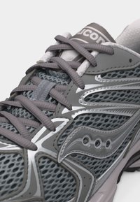 Γκρι και λευκό αθλητικό παπούτσι Saucony με επάνω μέρος από δίχτυ, συνθετικές επενδύσεις και γκρι κορδόνια που διασταυρώνονται σε λευκό φόντο.