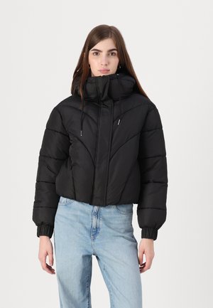 Sort pufferjakke med høj krave, snørede detaljer, quiltet tekstur, cropped design og elastiske manchetter. Parret med lyseblå jeans.