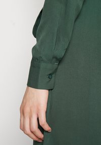 Prawa ręka spoczywa obok ciała, ubrana w ciemnozieloną długą rękawicę z zapięciem na guzik, na tle jasnym i jednolitym.