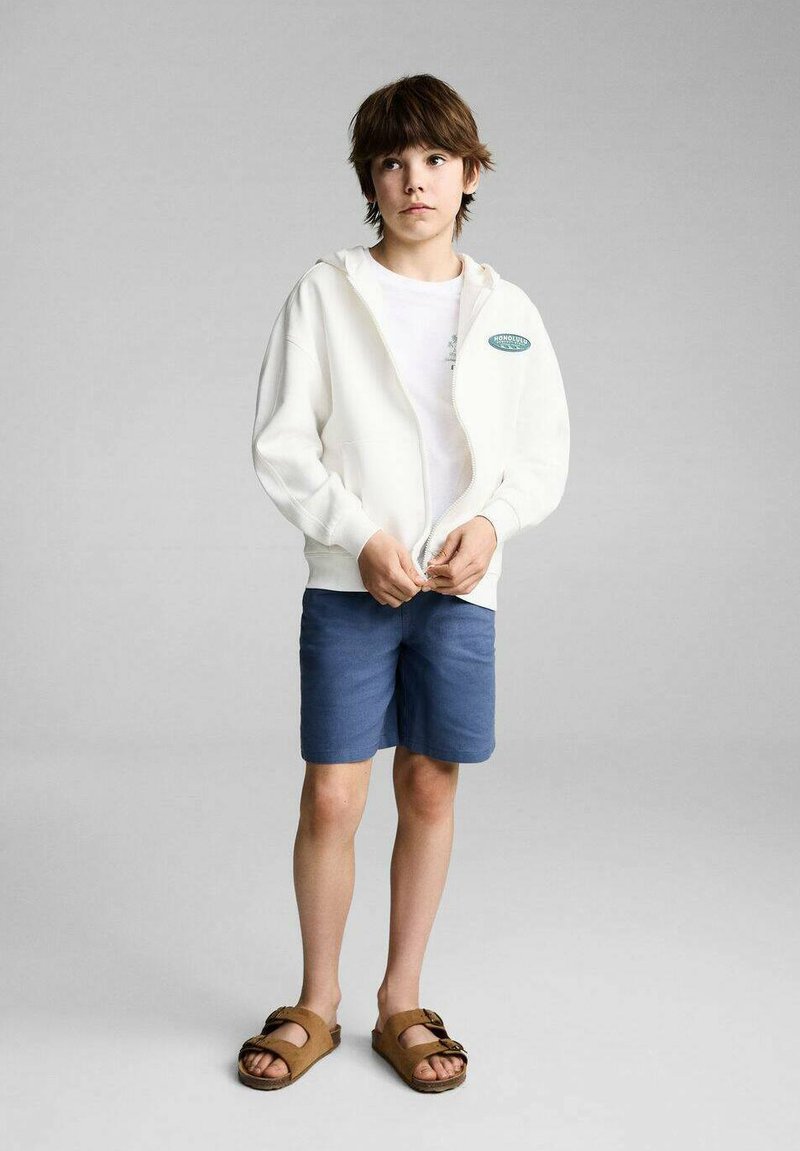 Felpa bianca con zip, t-shirt bianca sotto, pantaloni corti blu e sandali marroni. Design a taglio dritto, vestibilità rilassata e branding minimale sulla felpa.