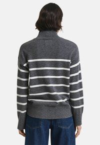 Pull gris à rayures horizontales blanches, avec un col montant et un ourlet côtelé. Texture douce et coupe décontractée. Porté avec un jean bleu foncé.