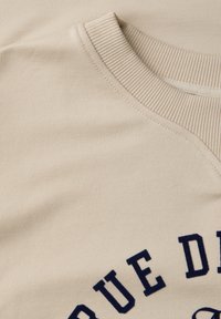 Sweatshirt beige avec un col côtelé texturé. Présente un texte bleu marine en arc de cercle sur le devant, fabriqué dans un tissu doux.
