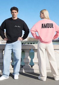 Homme en sweat-shirt noir « Mira Paris » et jeans clairs face à l’avant ; femme en sweat-shirt rose « AMOUR » et pantalon blanc tournée de dos près d’un pont sur la rivière.