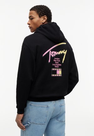 Άνδρας που φοράει μαύρη μπλούζα με κουκούλα με πολύχρωμο λογότυπο "Tommy" και ονόματα πόλεων στην πλάτη, σε συνδυασμό με ανοιχτόχρωμα μπλε τζιν, στραμμένος πλαγίως.