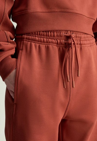 Zbliżenie na osobę w rdzawych spodniach dresowych z elastycznym pasem i zawiązanymi sznurkami, z ręką włożoną do lewego kieszeni.