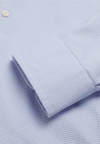 Cămașă de culoare albastru deschis cu un model în carouri mici. Prezintă un închidere cu nasture și manșetă pliată, realizată dintr-un material textil neted.