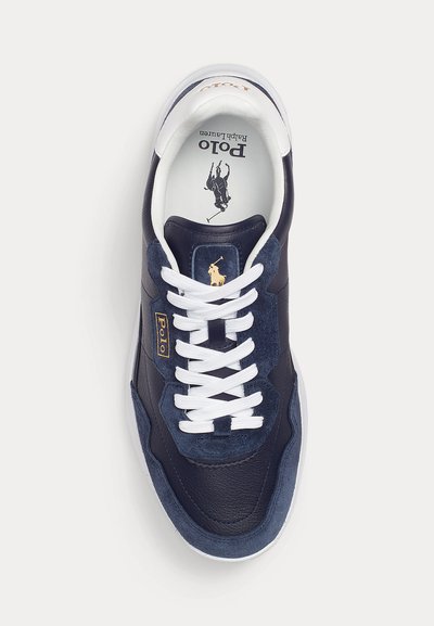 Tengeri kék sportcipő bőr és velúr felsőrésszel, fehér fűzőkkel, márkás nyelvvel és arany logóval. Texturált felület sima részletekkel.