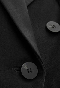 Gros plan sur le revers d'un manteau noir avec deux gros boutons noirs cousus sur un tissu en laine texturée.