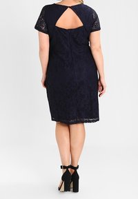Vestito in pizzo blu navy con maniche corte, caratterizzato da un design con schiena a occhio di chiave e una silhouette fino al ginocchio, abbinato a décolleté neri con cinturino alla caviglia.