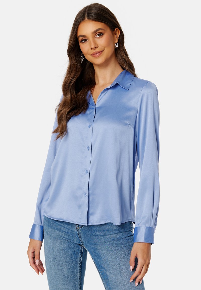 Blusa di seta azzurro chiaro con colletto, chiusura frontale a bottoni e maniche lunghe con dettagli ai polsini. Texture liscia, vestibilità comoda.