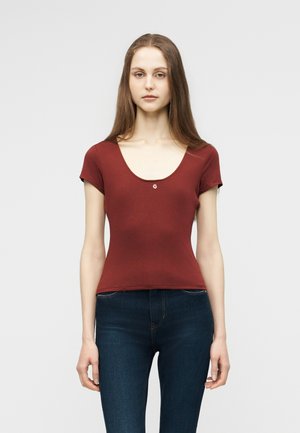 Γυναίκα με μακριά καστανά μαλλιά, που φοράει εφαρμοστό μπορντό μπλουζάκι με κοντά μανίκια και σκούρο μπλε τζιν, σε όρθια στάση μπροστά σε απλό ανοιχτό φόντο.