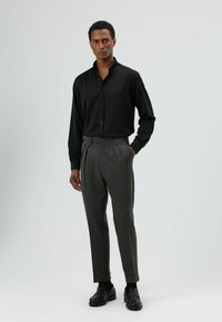 Zwarte longsleeve gecombineerd met grijze geplooide broek en zwarte loafers. Gladde textuur, nauwsluitende pasvorm en knoopdetails aan de broek.