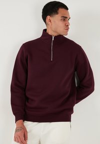 Sudadera burdeos con cremallera, fabricada en un tejido suave, que presenta un cuello alto y puños acanalados, con un diseño limpio y minimalista.