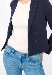 Granatowa marynarka z dwoma guzikami, noszona na jasnokremowej, falbaniastej bluzce oraz jasnoniebieskich, dopasowanych dżinsach. Prosty design z czystymi liniami.