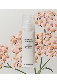 Biała cylindryczna butelka z przezroczystą pompką, oznaczona etykietą "Susanne Kaufmann Advanced Anti-Ageing Rich Cream", na różowym tle z kwiatami.