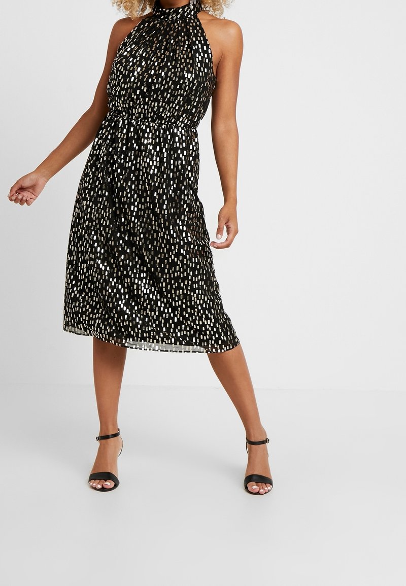 Žena v černých halenkových šatech po kolena se stříbrnými obdélníkovými vzory, stojící s roztaženýma nohama a v černých lodičkách s kotníkovým páskem na jednobarevném pozadí.