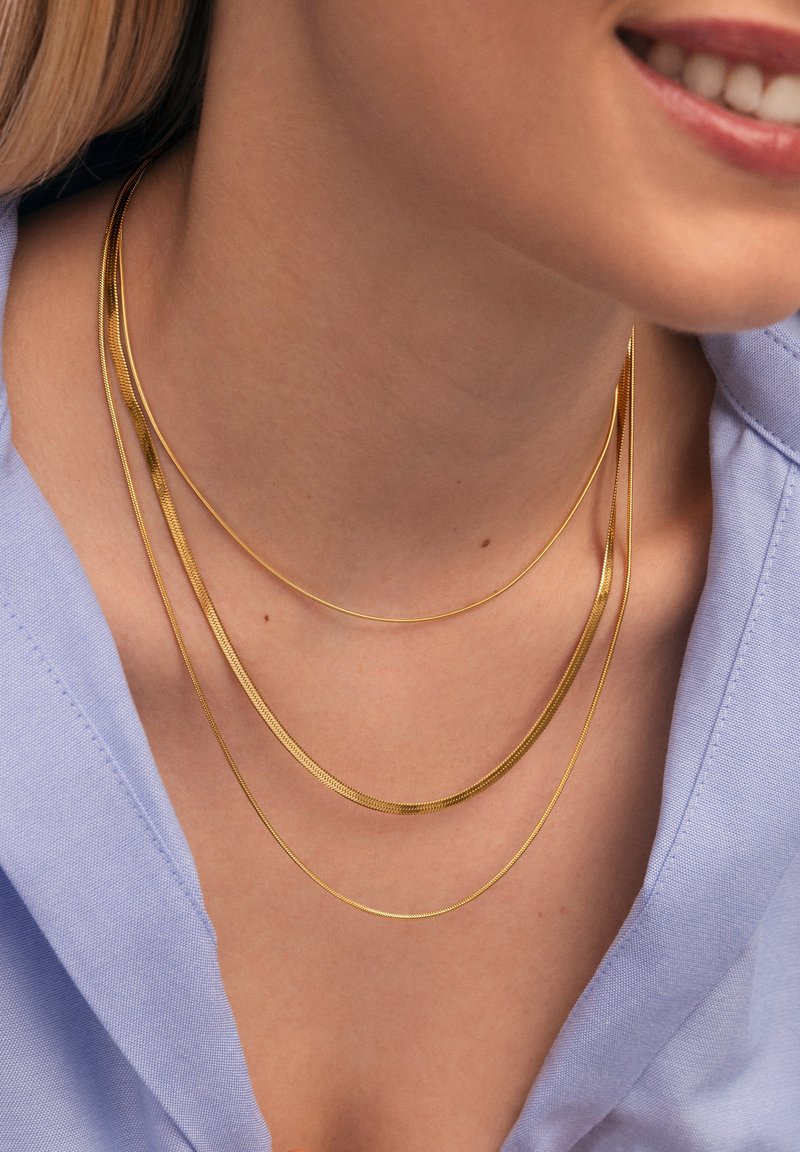 Tre delicati collier dorati di lunghezze diverse, con texture lisce e finitura lucida, sovrapposti a una camicia azzurro chiaro.