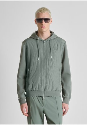 Giovane uomo con capelli biondi che indossa occhiali da sole scuri, una giacca con cappuccio verde chiaro con cerniera e pantaloni abbinati, in piedi contro uno sfondo semplice.