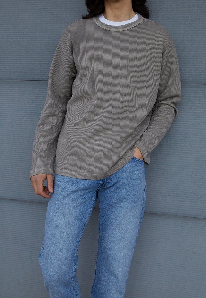 Ljusgrå räfflad långärmad skjorta med rundad krage, lager på lager över en vit T-shirt med rund hals, tillsammans med blå raka jeans.