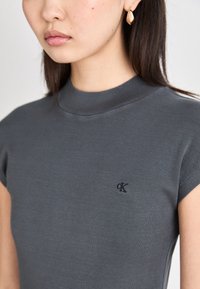 Γκρι, κοντομάνικο τοπ με ψεύτικο γιακά, που διαθέτει ένα μικρό μαύρο λογότυπο στο στήθος. Κατασκευασμένο από ένα λείο, εφαρμοστό υλικό.