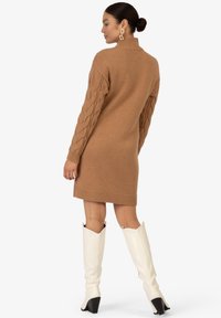 Kamel gefärbtes Strickkleid mit hohem Ausschnitt und Kabelstrickärmeln, kombiniert mit weißen High-Heels und groben goldenen Creolen.