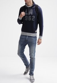 Felpa con cappuccio blu navy con accenti grigi, grande stampa "LA 1962", abbinata a jeans azzurri chiari e sneakers grigie, indossata con uno zaino nero.