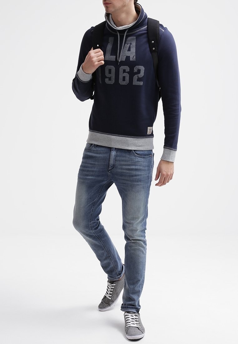 Felpa con cappuccio blu navy con accenti grigi, grande stampa "LA 1962", abbinata a jeans azzurri chiari e sneakers grigie, indossata con uno zaino nero.