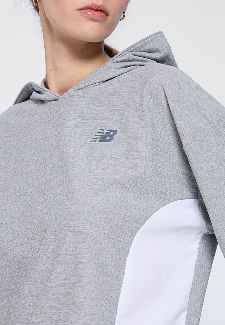 Sudadera gris clara de tejido suave, con un panel lateral blanco y un pequeño logo gris en el pecho. Diseño con capucha y ajuste relajado.