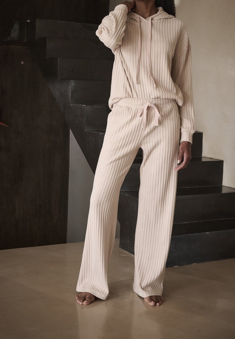 Beżowy dres z prążkowanego materiału składający się z bluzy z kapturem i długimi rękawami oraz spodni z gumką w pasie, wykonany z fakturowanej tkaniny o luźnym kroju.