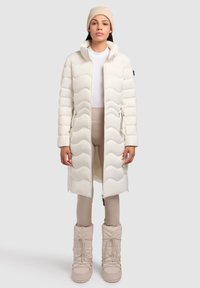 Geacă puffer albă, matlasată, cu guler înalt și buzunare cu fermoar, asociată cu cizme bej izolate și pantaloni bej ajustați.