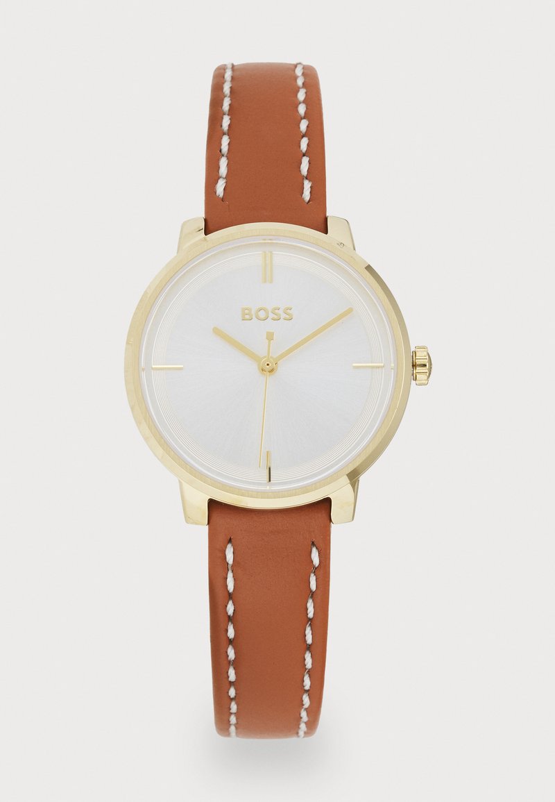 Reloj de tono dorado con una esfera blanca. La correa de piel marrón presenta costuras blancas. Caja redonda sencilla y marcadores de horas minimalistas.