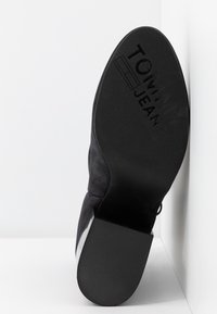 Botín negro de ante hasta el tobillo con suela de goma texturizada, presenta el logo en relieve "TOMMY JEANS", vista en ángulo que resalta el talón y el material.