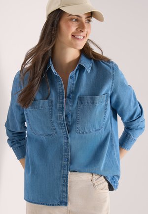 Kobieta uśmiechająca się, w beżowej czapce, niebieskiej dżinsowej koszuli zapinanej na guziki z dwoma kieszeniami na piersi oraz jasnobeżowych spodniach, stojąca z rękami za plecami.