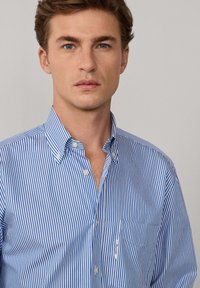 Camisa de botones a rayas azul y blanca con cuello, bolsillo en el pecho y textura de tela ligera. Presenta botones blancos y una pequeña etiqueta.