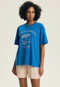 Kobieta ubrana w niebieski oversize'owy t-shirt z napisem "Costa D'Amalfi" i grafiką plaży, w połączeniu z jasnoróżowymi szortami w paski.