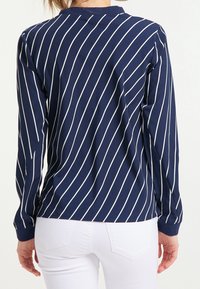 Marineblaues Langarmshirt mit weißen vertikalen Streifen, rundem Ausschnitt und strukturiertem Stoff. Weiße Jeans sind am unteren Ende sichtbar.