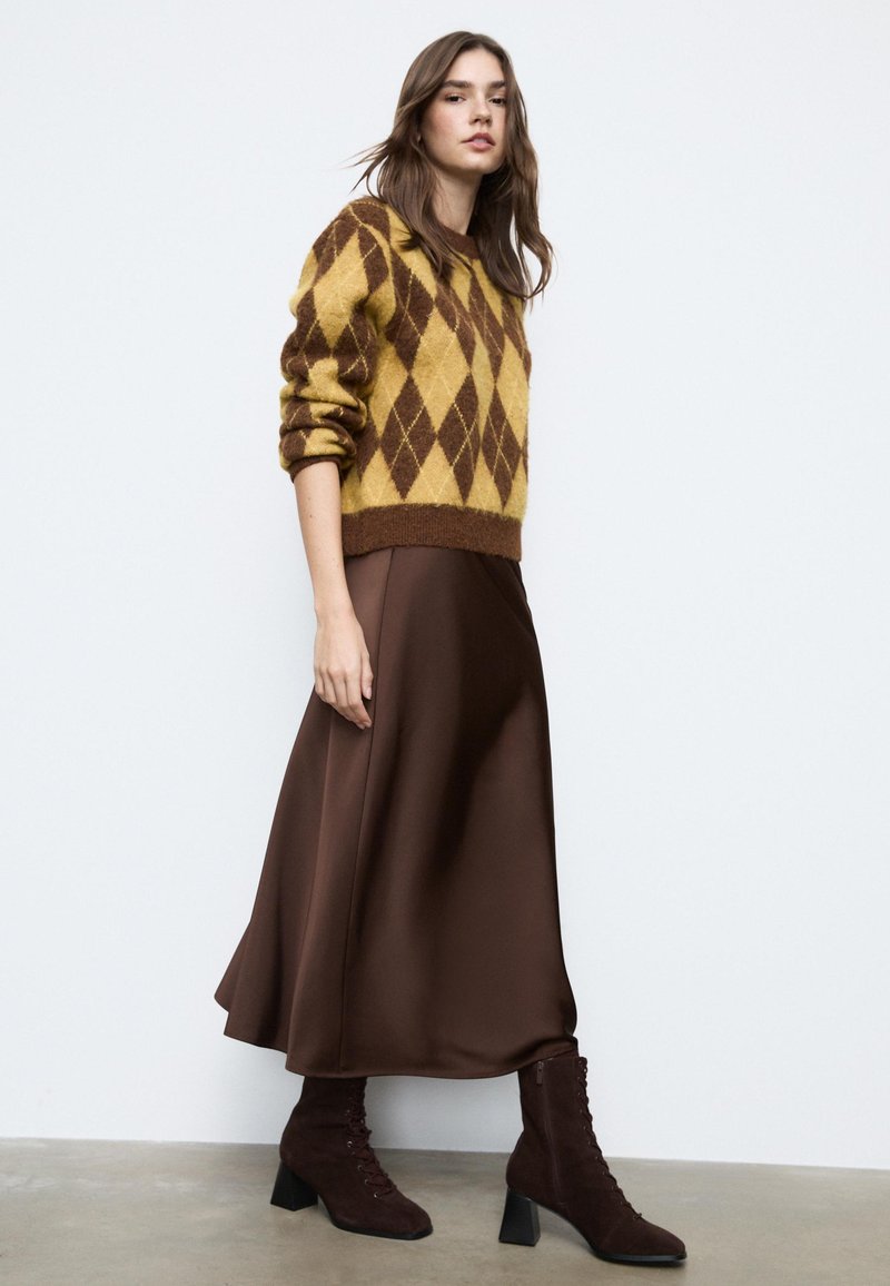 Hnědá saténová sukně s vysokým pasem v kombinaci s žlutým a hnědým svetr s argyle vzorem a tmavě hnědými šněrovacími kotníkovými botami.