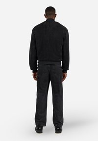 Giubbotto in shearling nero con colletto alto e polsini rigati, abbinato a jeans scuri e larghi e sneakers nere, visto da dietro.