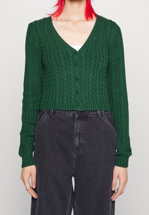 Personne portant un cardigan vert boutonné en maille torsadée par-dessus un haut blanc, associé à un jean taille haute gris foncé, visible du cou aux hanches.