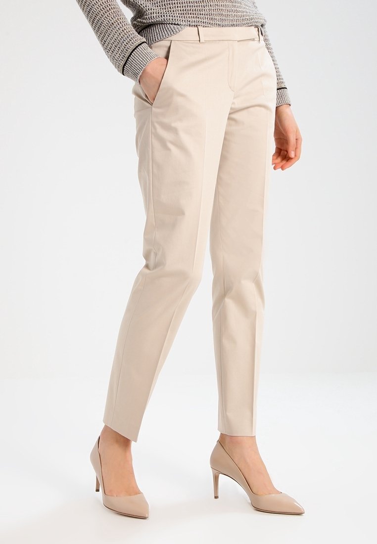 Pantalon beige sur mesure avec une texture lisse, doté de poches latérales et d'une coupe effilée, complété par des talons hauts de couleur neutre.