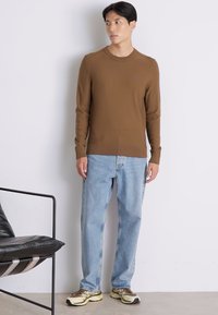 Brauner, gestrickter Pullover mit langen Ärmeln, kombiniert mit hellblauen Jeans und beigen Turnschuhen mit dunklen Akzenten. Stehend vor einem neutralen Hintergrund.