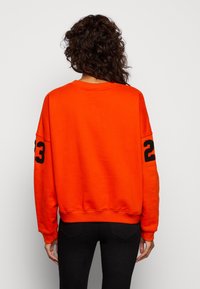Sudadera naranja con cuello redondo y el número "23" en negro en la manga. Hecha de material suave, presenta un corte holgado y un dobladillo de canalé.