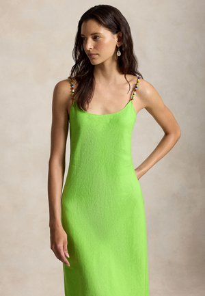 Kvinde med langt mørkt hår iført en ærmeløs, skriggrøn kjole med flerfarvede perlestraps, stående mod en ensfarvet lys baggrund.