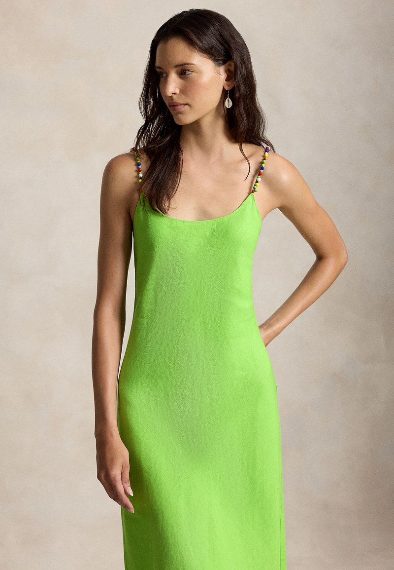 Hosszú sötét hajú nő, élénk zöld, ujjatlan ruhában, amelynek színes gyöngyös pántjai vannak, egyszínű világos háttér előtt áll.