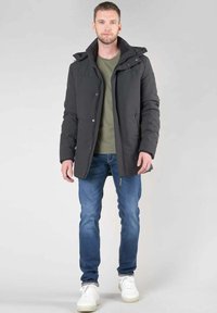 Zwarte gewatteerde jas met capuchon, gecombineerd met een groen T-shirt en blauwe spijkerbroek. Opvallende details zijn de voorzakken en ritssluitingen.