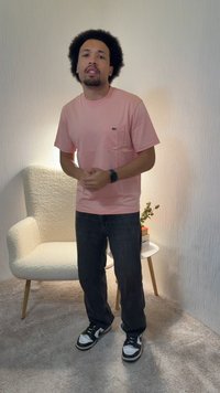 Άνδρας με σγουρά μαλλιά, ροζ πουκάμισο και μαύρο παντελόνι, στέκεται σε δωμάτιο με χαλί κοντά σε λευκή πολυθρόνα και μικρό τραπέζι με βιβλία και φυτό.