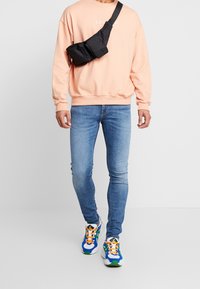Ljusrosa sweatshirt med logotyp, svart axelväska, ljusblå jeans och färgglada sneakers med blå, grön, orange och vita detaljer.