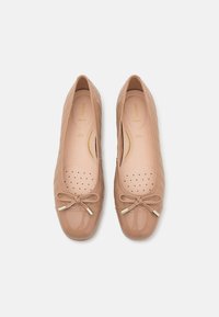 Paire de ballerines matelassées beiges avec petits nœuds et bouts ronds brillants, vues de dessus sur un fond uni.