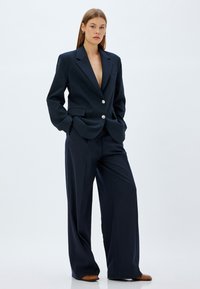 Blazer entallado azul marino con escote en V profundo, cierre de un botón y bolsillos laterales, combinado con pantalones de pierna ancha a rayas.